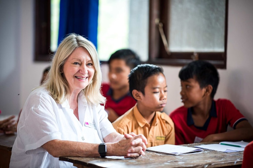 A blond woman sits next to two Balinese boys at a school desk.