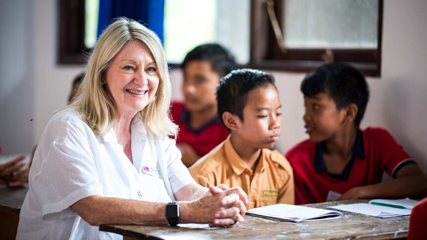 A blond woman sits next to two Balinese boys at a school desk.
