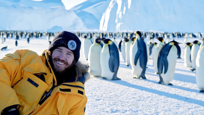 Smiling man surrounded by emperor penguins in Antarctica in a story about working as a tradie.