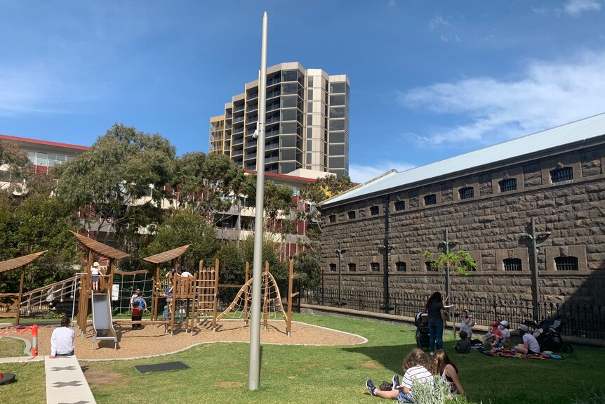 A playground with children and families around it, near bluestone Pentridge walls, with tall apartment block in background.