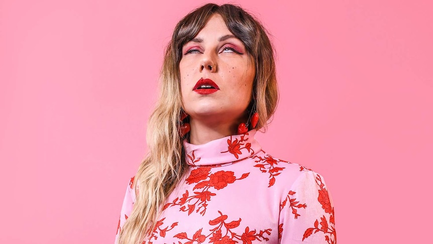 Photograph of Maddy Jane rolling her eyes, wearing a pink and red dress infront of a pink background