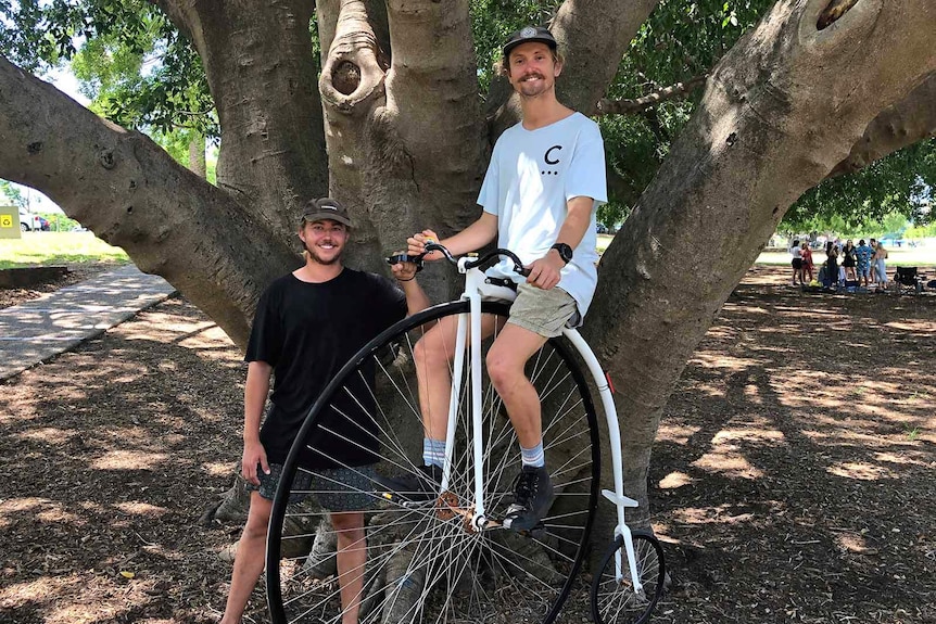 A man smiling atop a penny farthing bicycle, supported by a friend standing beside him