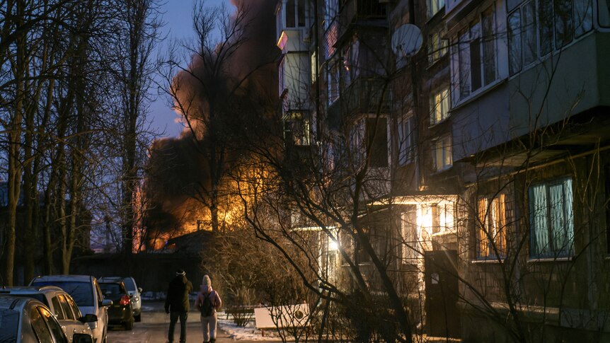 Local residents walk near a residential building as infrastructure burns.