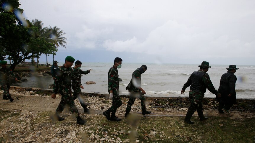 On an overcast day Indonesian military officers walk past a coastline with a high tide searching for tsunami survivors.