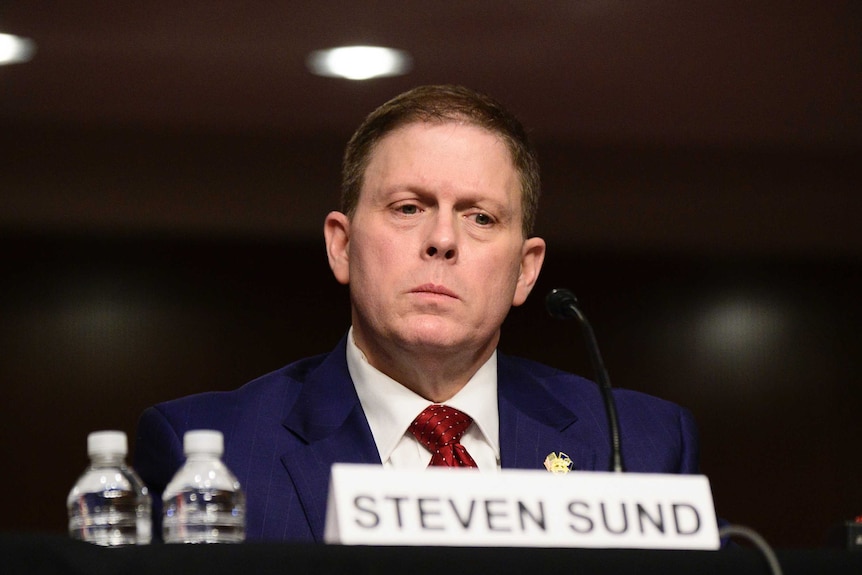 A man in a suit looks downward as he testifies from behind a small microphone.