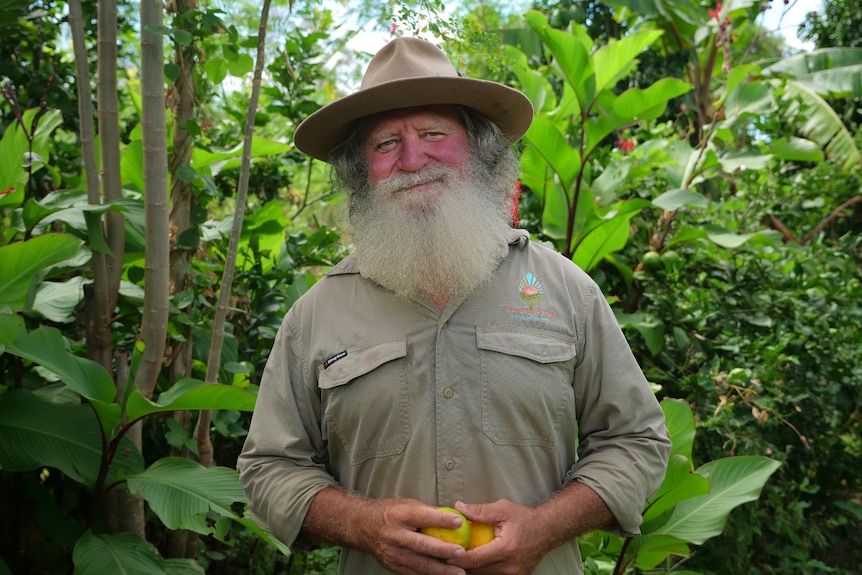 Ross holding fruit, smiling at the camera he has a large beard, wide-brimmed hat, greenery surrounds him.
