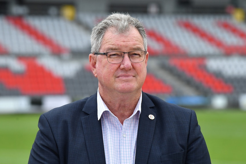 A man with short grey hair and glasses stands on a sports field wearing a suit.