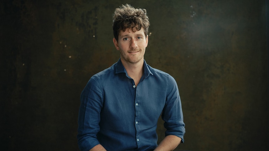 A white man with curly light brown hair and blue button up shirt looks at the camera with his hands clasped