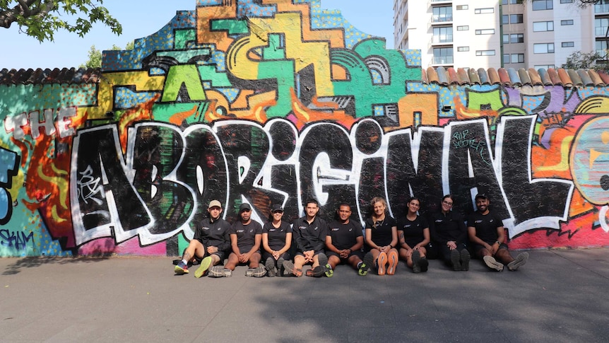 A group of young men and women sit on the ground in front of a graffiti wall with the words "Aboriginal" painted on it.