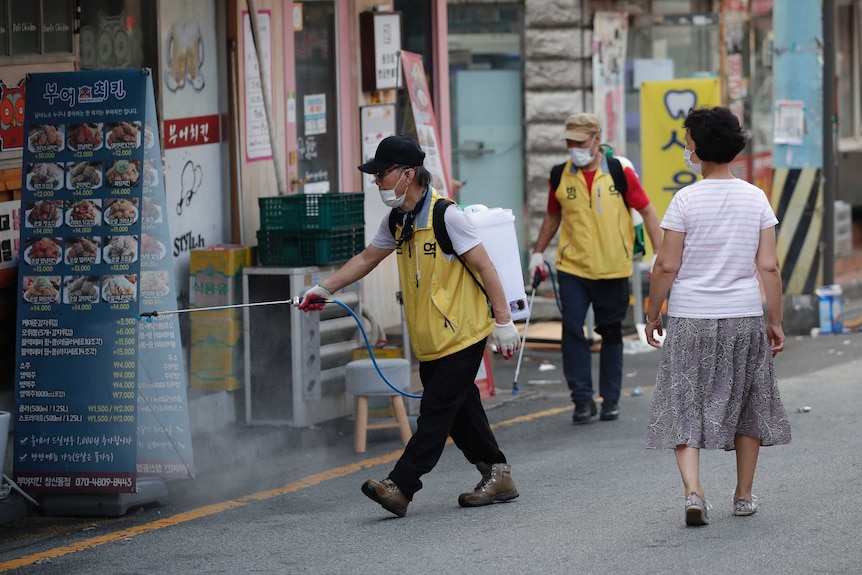 A worker in a yellow vest uses a spraying device to disinfect food signs along the side of s street.