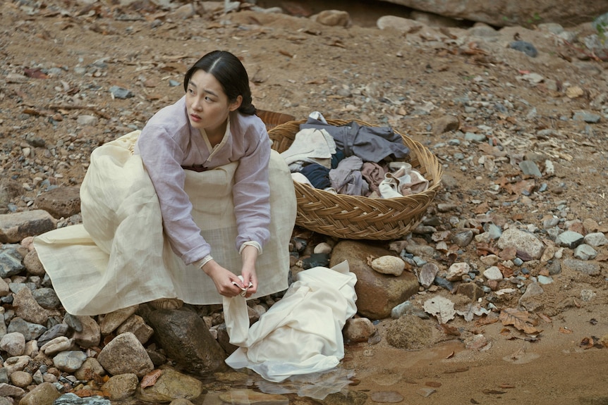 A young woman in traditional Korean clothing looks up from doing her laundry by a river.