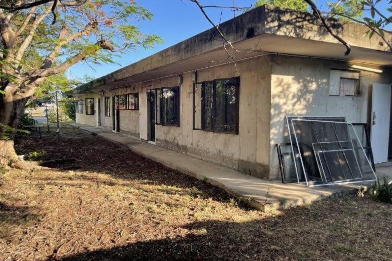 The side of a fully concrete house with a dirt garden and some fly screens leaning against it.