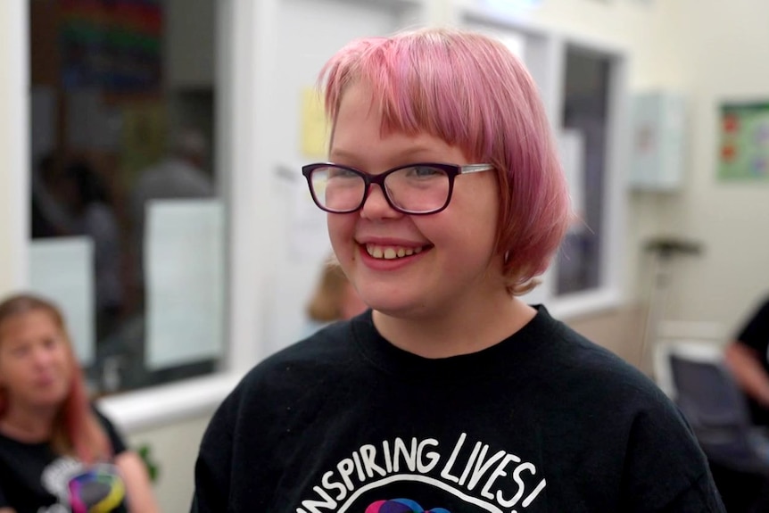 A photo of a 12-year-old girl smiling with pink hair and glasses.