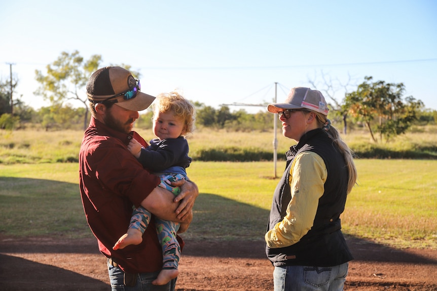 A man holds a toddler on his hip as he speaks to a woman in the sunshine