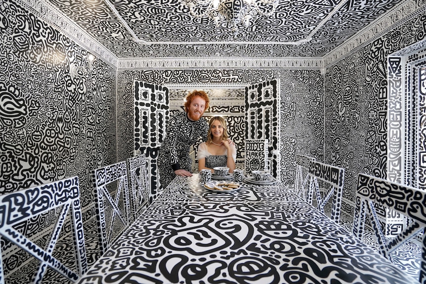 Sam Cox and his wife at the end of a dinner table. The room, including their clothes, is covered in black and white art.