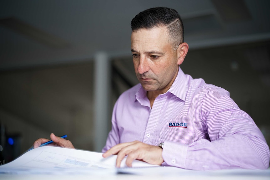 A man with short brown hear, wearing a purple shirt with Badge written on it looks at building planning documents.