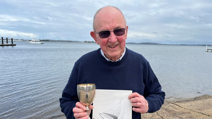 An elderly man holds a silver trophy with lake in background. 