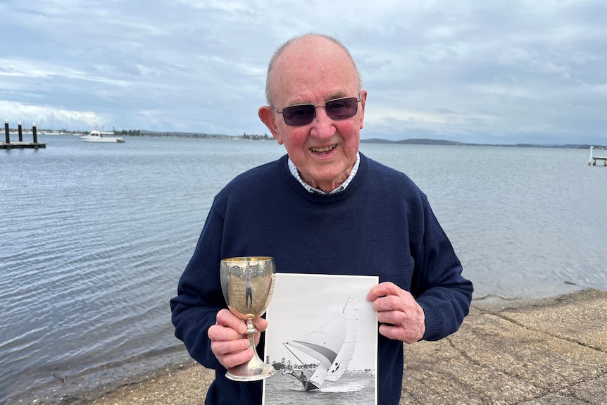 An elderly man holds a silver trophy with lake in background. 