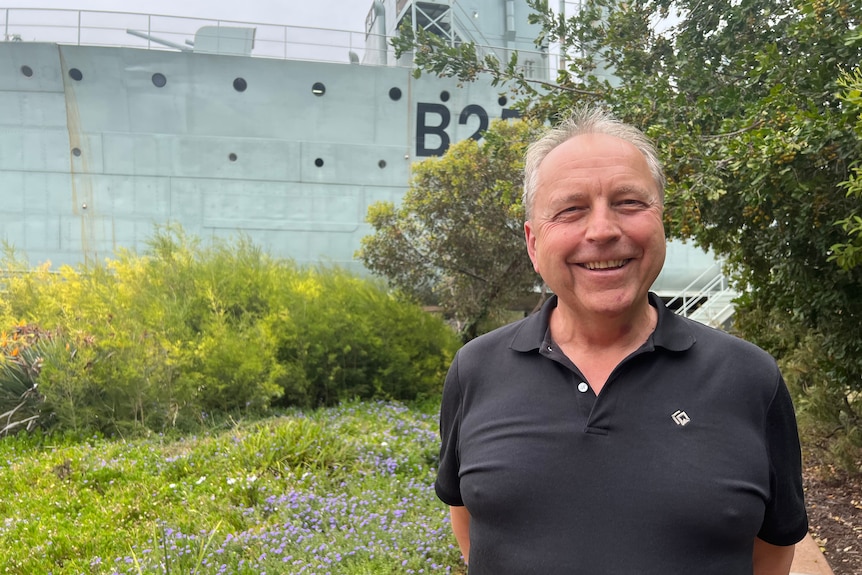 A man smiling, standing in front of an old warship that is part of the Whyalla Maritime Museum. 
