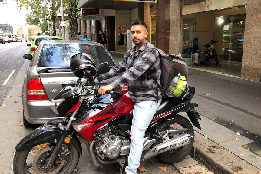 A man in a shirt and jeans wears a food delivery bag and sits on a red and black motorcycle that is parked on the street.