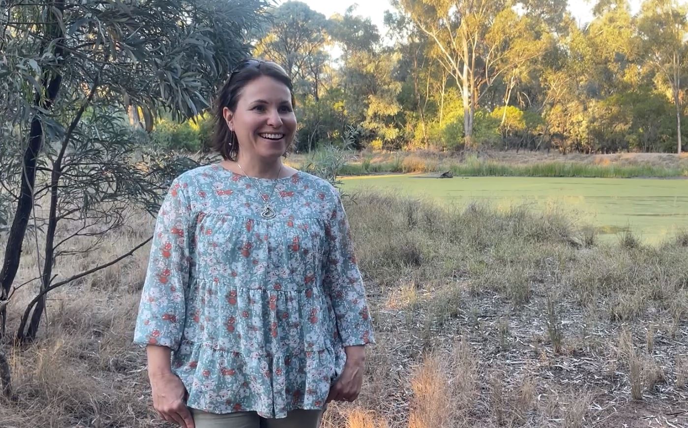 A woman with dark hair stands in a bushy area, smiling.