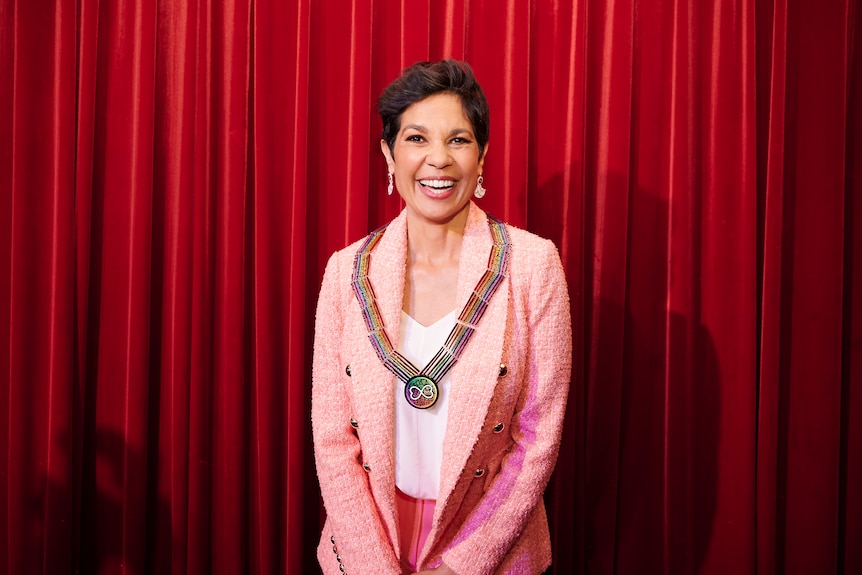 A dark-haired Indigenous woman in a white shirt and pale pink jacket, smiles widely as she wears her WorldPride medallion