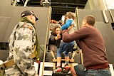 A child is seen being lifted by a man into the arms of a woman aboard MV Sycamore. A Naval officer stands to the left, watching.