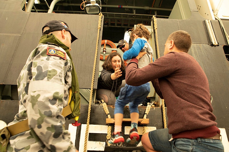 A child is seen being lifted by a man into the arms of a woman aboard MV Sycamore. A Naval officer stands to the left, watching.