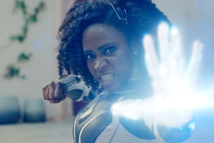 A black woman in her 20s wearing a superhero suit takes a strong stance and shoots a blue beam of electricity from her hand.