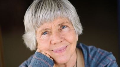 A woman with grey hair and blue jumper and tan shirt smiles with mouth closed.