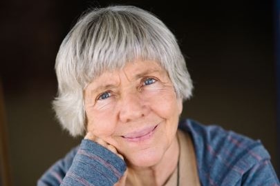 A woman with grey hair and blue jumper and tan shirt smiles with mouth closed.