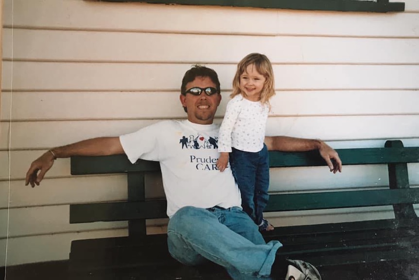 Raina Collett as a child stands smiling next to her dad, Steve, who is sitting on a wooden bench.
