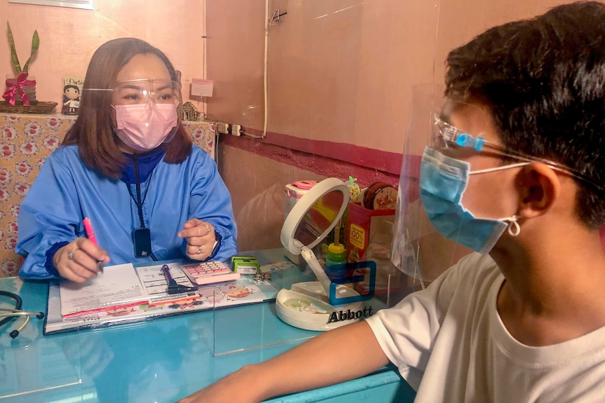A young Filipina woman in a face mask and scrubs talks to a boy across a table