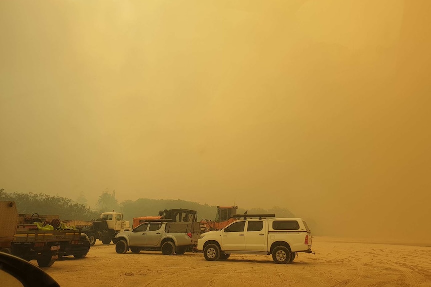 Orange smoke haze on the beach with cars and a bulldozer parked.