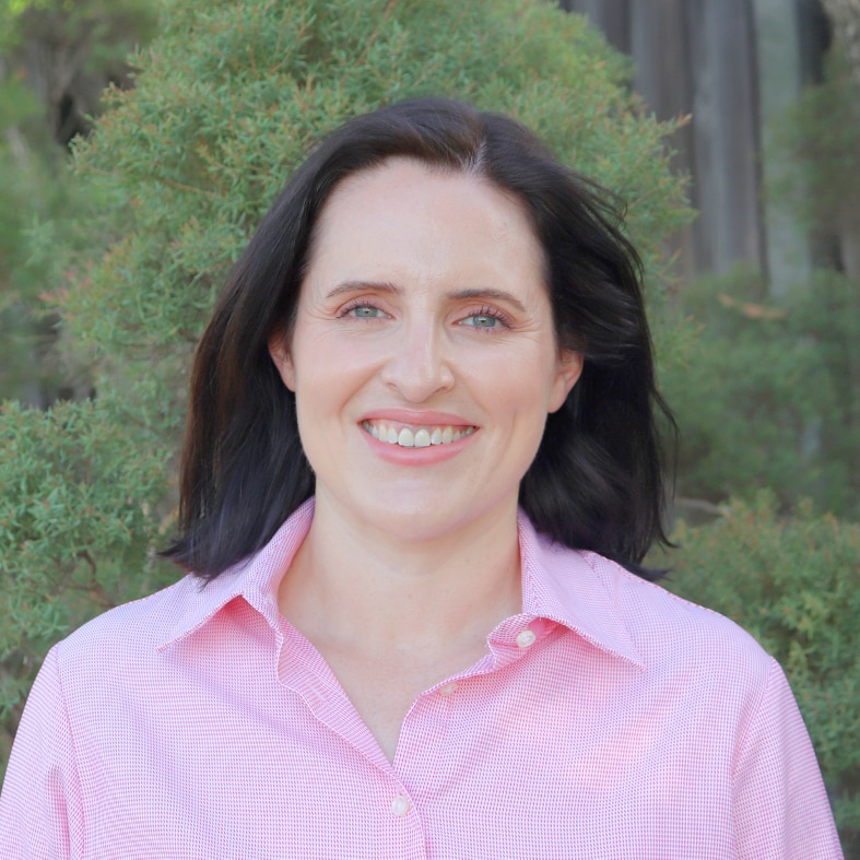 Woman in button up shirt, smiling in front of green foliage