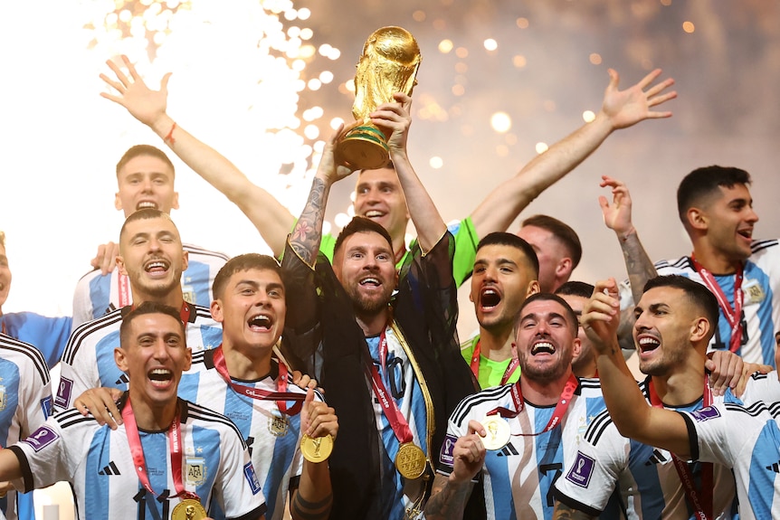 A smiling man lifts a golden trophy among celebrating teammates with fireworks exploding in the background.