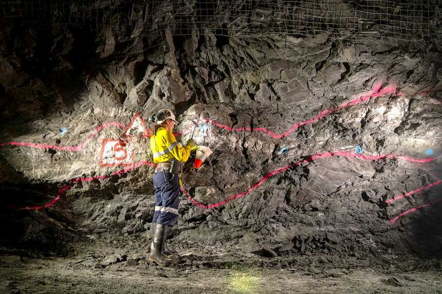 A woman in hi-vis clothing standing in a cave with a torch undergound at Bellevue Gold mine