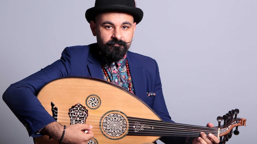 Joseph Tawadros sits in front of a grey backdrop holding an oud and looks at the camera.