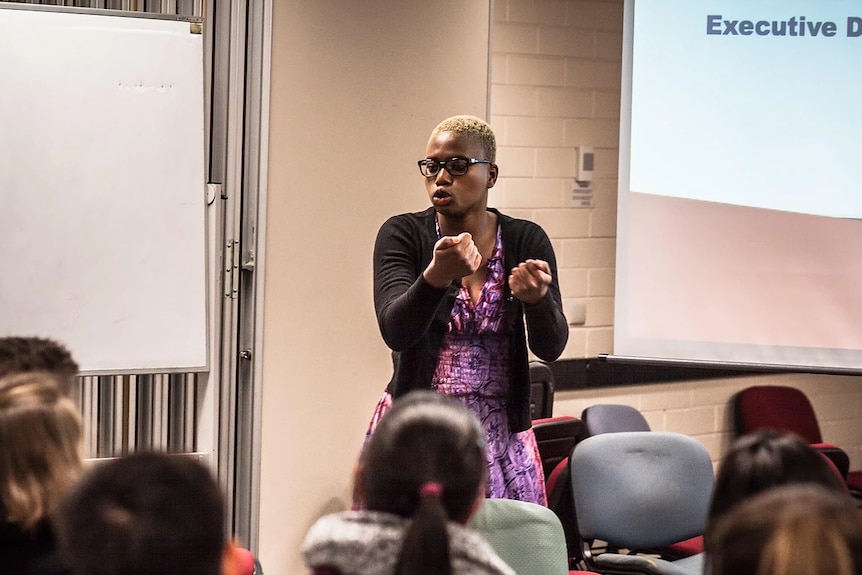 Woman wearing a purple dress and glasses addresses a room full of young medical students