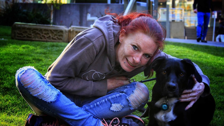 Bek Eirth sits cross legged on the grass hugging her dog Dexter who is beside her.