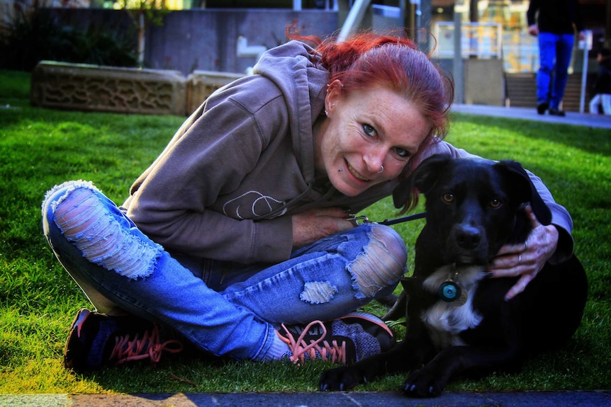 Bek Eirth sits cross legged on the grass hugging her dog Dexter who is beside her.