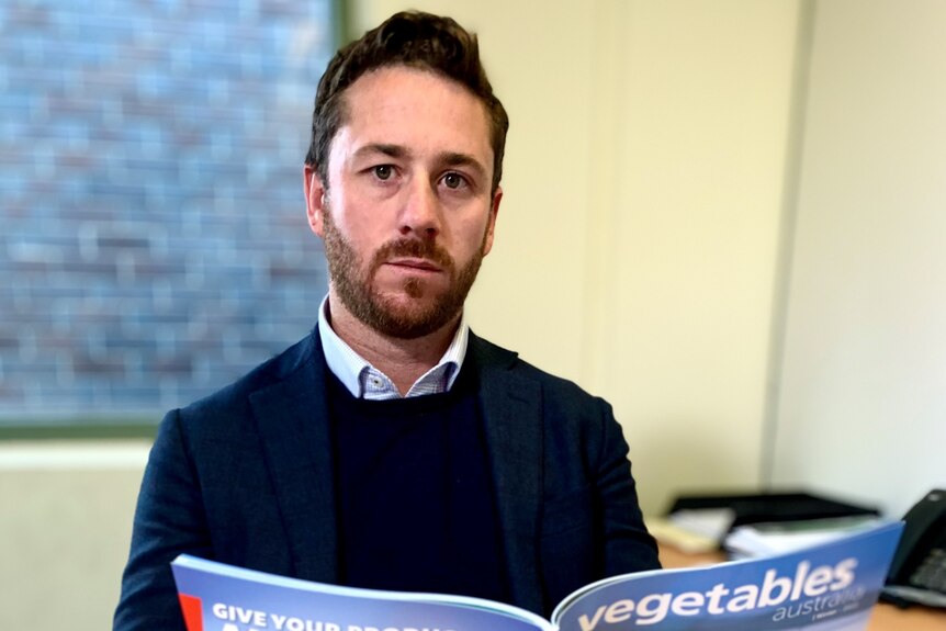 A young man, neat beard, wearing sweater over shirt, reads a magazine with vegetables written on the cover. 