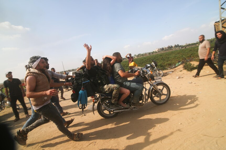A woman sits between two men on a motorcycle covering her head as they drive away, three more men run behind them as others look