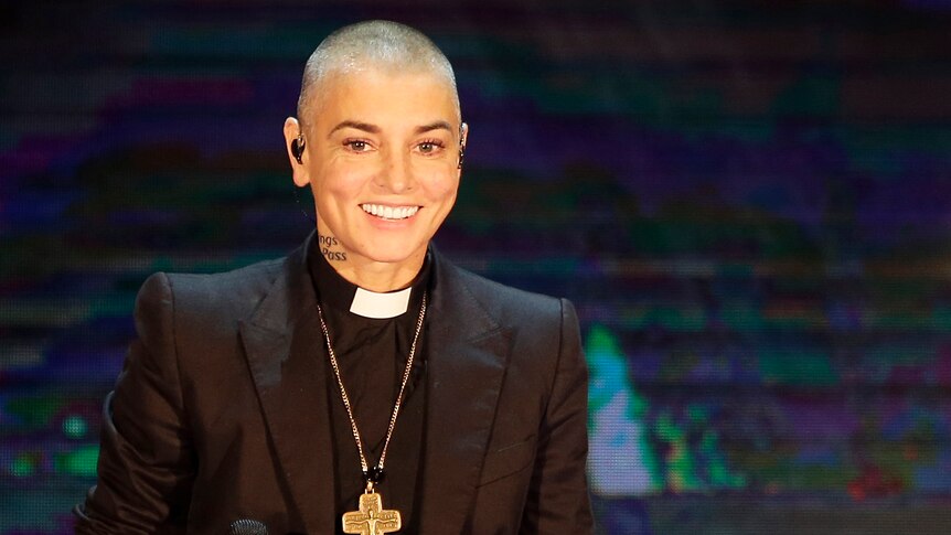 Sinead O'connor wearing a priests black outfit with a cross holding a microphone and smiling