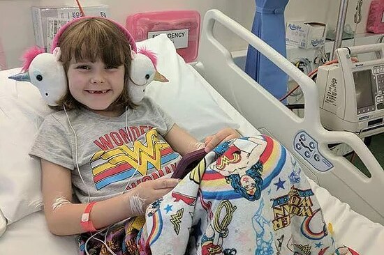 Freyja sits in a hospital bed, smiling, with a tablet computer on her lap and headphones on her ears.