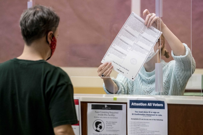 An election official behind a plastic barrier holds up a large ballot paper to a voter. Both are wearing masks.