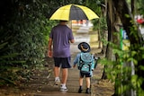 A man holds an umbrella while walking with a primary school-aged child in the rain on a street.