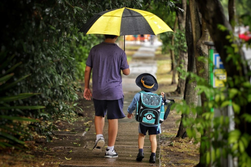 A man holds an umbrella while walking with a primary school-aged child in the rain on a street.