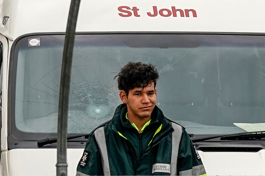 A St John Ambulance worker leans on the bonnet of an ambulance that has a smashed windscreen.