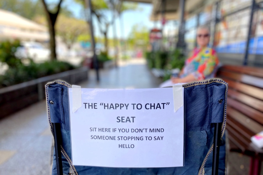 The back of a camping chair on a main street with a notice stuck to it that reads, "The Happy to Chat Seat".